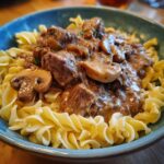 A close-up of a bowl of Beef Stroganoff, featuring tender beef chunks and mushrooms in a creamy sauce served over egg noodles.