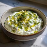 A close-up of a bowl of creamy Tzatziki sauce topped with fresh dill, lemon zest, and a drizzle of olive oil.