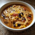 A close-up of a white bowl filled with hearty Crockpot Chicken Enchilada Soup, topped with shredded chicken, black beans, corn, and cheese.