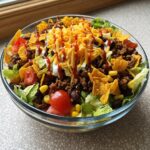 A large glass bowl filled with Dorito Taco Salad, featuring ground beef, lettuce, tomatoes, black beans, corn, shredded cheese, and crushed Doritos.