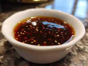 Close-up of a white dipping bowl filled with a rich, dark Dumpling Sauce, speckled with sesame seeds and red chili flakes.