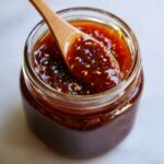 A wooden spoon scoops out thick, glossy honey BBQ sauce from a glass jar, sprinkled with sesame seeds.