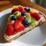 A slice of fruit pizza sugar cookie topped with cream cheese frosting and fresh strawberries, blueberries, kiwi, and orange slices.
