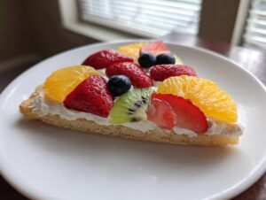 A slice of fruit pizza sugar cookie topped with cream cheese frosting and fresh strawberries, blueberries, kiwi, and orange slices.