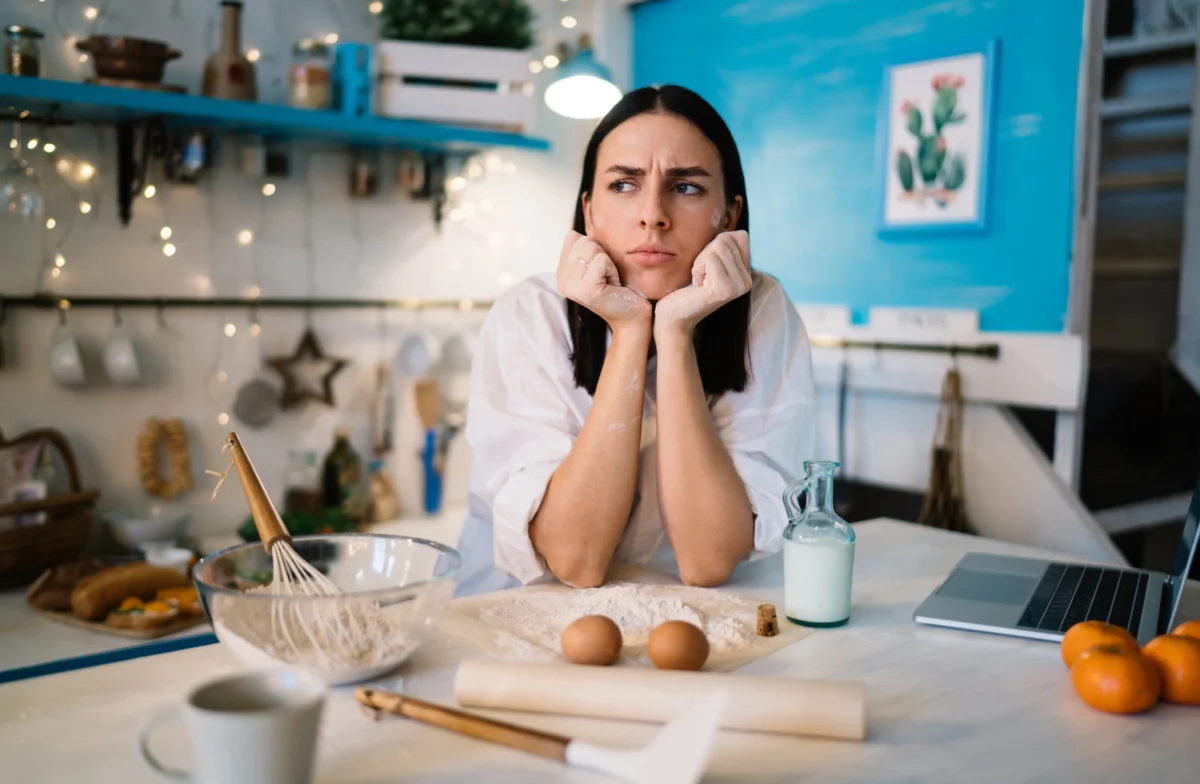 Maddie Thompson with a thoughtful expression, planning a recipe in her kitchen with flour, eggs, and a laptop nearby.