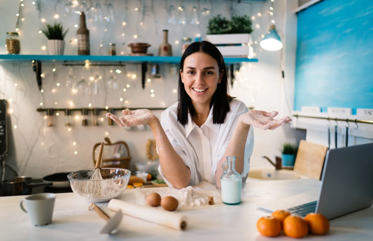 Maddie Thompson smiling warmly at the camera with her hands open, gesturing to a kitchen counter filled with simple baking ingredients.