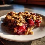 A close-up of a slice of sour cherry crisp with a golden oat topping and juicy cherries on a white plate.