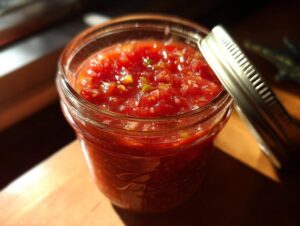 Close-up of a glass jar filled with vibrant homemade tomato salsa, with its lid resting beside it. This is a tomato salsa canning recipe.