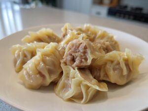 A close-up of several Traditional Chinese Dumplings on a white plate, one is cut open showing the savory pork filling.