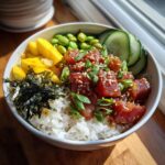 A vibrant Tuna Poke Bowl with fresh tuna cubes, rice, mango, edamame, cucumber slices, and seaweed, garnished with sesame seeds and green onions.