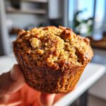 A close-up shot of a hand holding a golden-brown apple cinnamon muffin with visible spices and a nut on top.