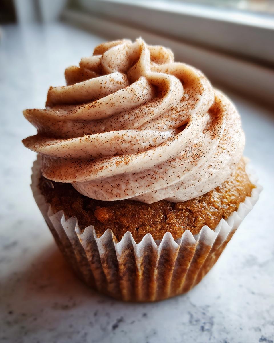 A close-up of a single apple pie cupcake topped with swirled cinnamon frosting and a sprinkle of cinnamon.
