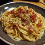 A close-up of creamy bacon carbonara pasta topped with crispy bacon bits and black pepper.