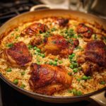 Close-up of a skillet filled with golden brown baked chicken and rice, garnished with peas and parsley.