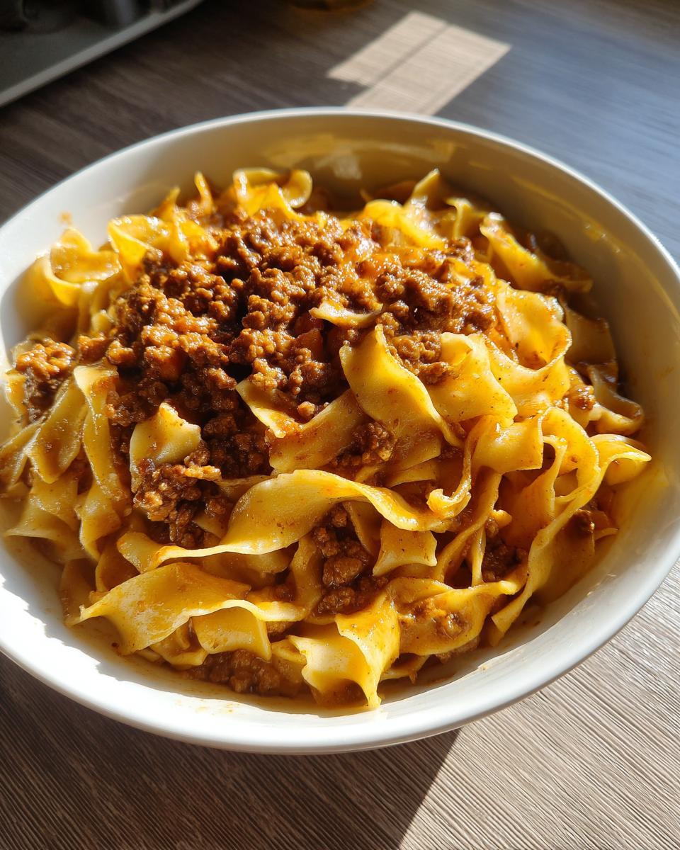 Close-up of a white bowl filled with delicious beef and noodles, topped with a rich meat sauce.