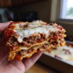 A close-up shot of a thick slice of beef lasagna, showing layers of pasta, rich meat sauce, and melted cheese.