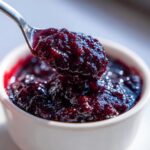 A close-up shot of a spoonful of glistening homemade blueberry jam being lifted from a white bowl.