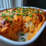 A close-up of a bubbling classic buffalo chicken dip in a white baking dish, topped with melted cheese and green onions.