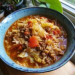 A close-up of a steaming blue bowl filled with cabbage roll soup, featuring ground beef, rice, cabbage, and tomatoes.