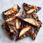 Close-up of broken pieces of dark chocolate caramel apple bark topped with apple slices and sea salt.