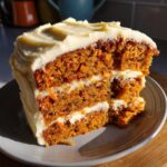 A thick slice of carrot cake with generous carrot cake cream cheese frosting, showing two layers of cake and frosting.