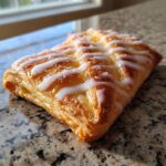 A close-up of a golden brown cheese danish, drizzled with white icing and sprinkled with coarse sugar.