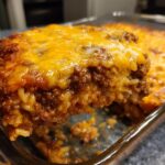 A close-up view of a slice of cheesy hamburger rice casserole in a baking dish, showing layers of ground beef, rice, and melted cheese.