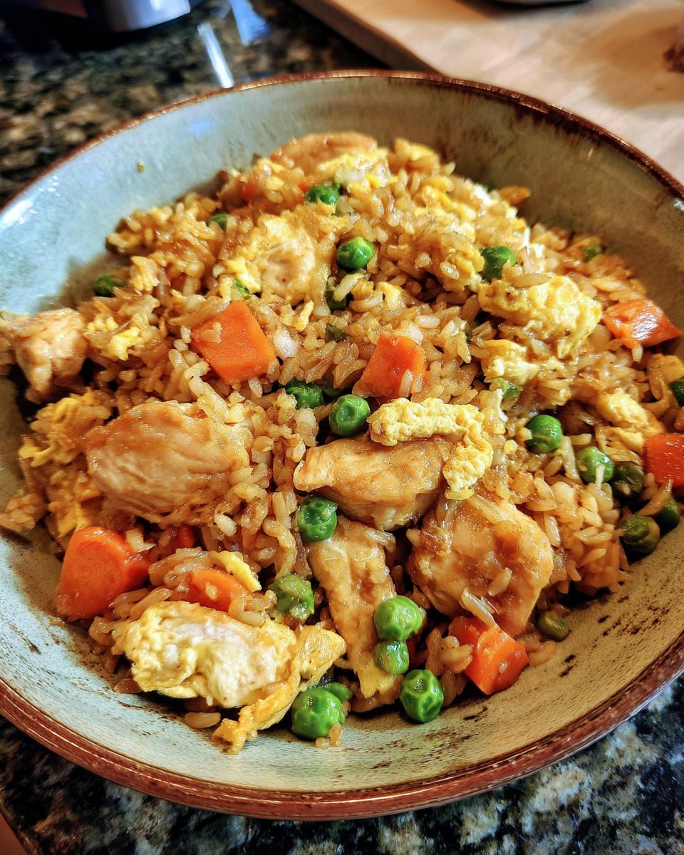 A close-up of a bowl filled with delicious chicken fried rice, featuring tender chicken pieces, peas, carrots, and scrambled egg.