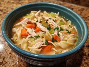 A close-up of a steaming bowl of chicken noodle soup, featuring shredded chicken, wide egg noodles, carrots, celery, and parsley.