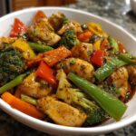 A close-up shot of a white bowl filled with delicious chicken stir fry with vegetables, including broccoli, carrots, bell peppers, and green beans.