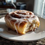 A close-up of a freshly baked cinnamon roll with a generous cream cheese glaze, on a white plate.