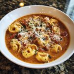 A close-up of a white bowl filled with crockpot tomato tortellini soup, topped with shredded cheese and parsley.