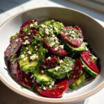 A close-up of a vibrant cucumber beetroot salad with crumbled feta cheese and fresh herbs in a bowl.