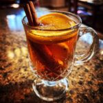 A clear glass mug filled with warm Dutch oven apple cider, garnished with orange slices and cinnamon sticks.