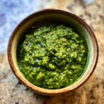 A close-up overhead view of vibrant green basil pesto in a rustic brown bowl.