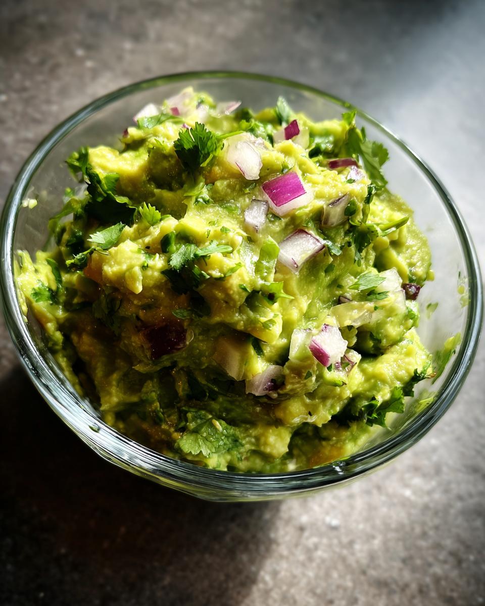 Close-up of fresh guacamole in a clear glass bowl, topped with red onion and cilantro.