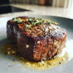 A perfectly cooked garlic butter steak, seasoned with herbs and resting in its own juices on a plate.