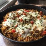 A close-up of a one-pot ground beef orzo dish in a cast iron skillet, topped with melted cheese, diced tomatoes, and parsley.