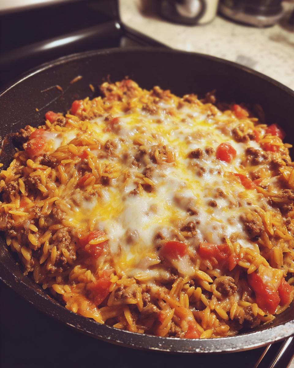 A close-up of a skillet filled with ground beef orzo pasta, diced tomatoes, and melted cheese.
