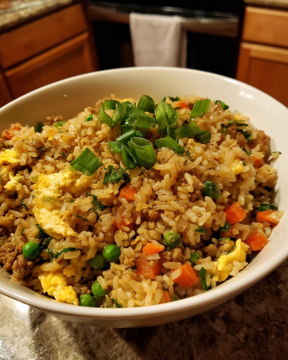 A close-up of a white bowl filled with savory ground chicken fried rice, featuring peas, carrots, scrambled egg, and green onions.