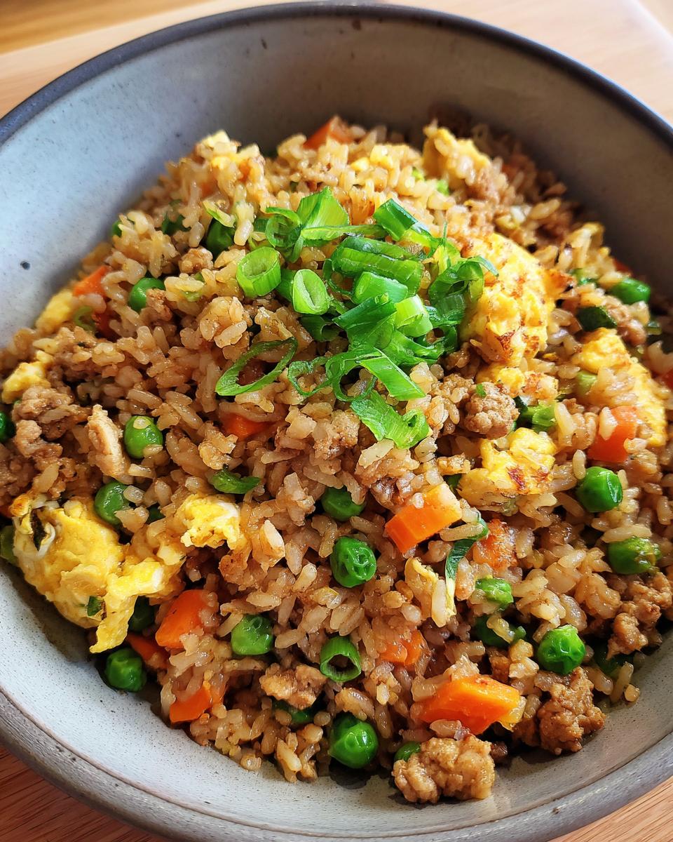 A close-up overhead view of a bowl filled with flavorful ground chicken fried rice, featuring peas, carrots, scrambled egg, and green onions.