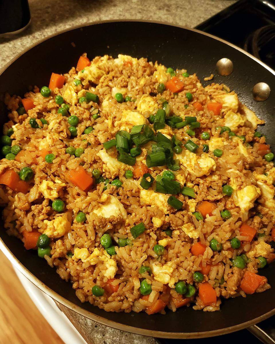 A close-up overhead view of delicious ground chicken fried rice in a pan, featuring peas, carrots, scrambled egg, and green onions.
