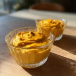 Two clear glass bowls filled with creamy, vibrant orange high protein pumpkin mousse on a wooden table.