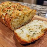 A close-up of a loaf of Italian herb and cheese bread, with one slice cut and showing herbs and melted butter.