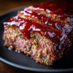 Close-up of sliced meatloaf with a rich, glossy glaze on top, served on a black plate.
