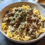 A close-up of a bowl of old fashioned beef stroganoff served over wide egg noodles, garnished with fresh parsley.