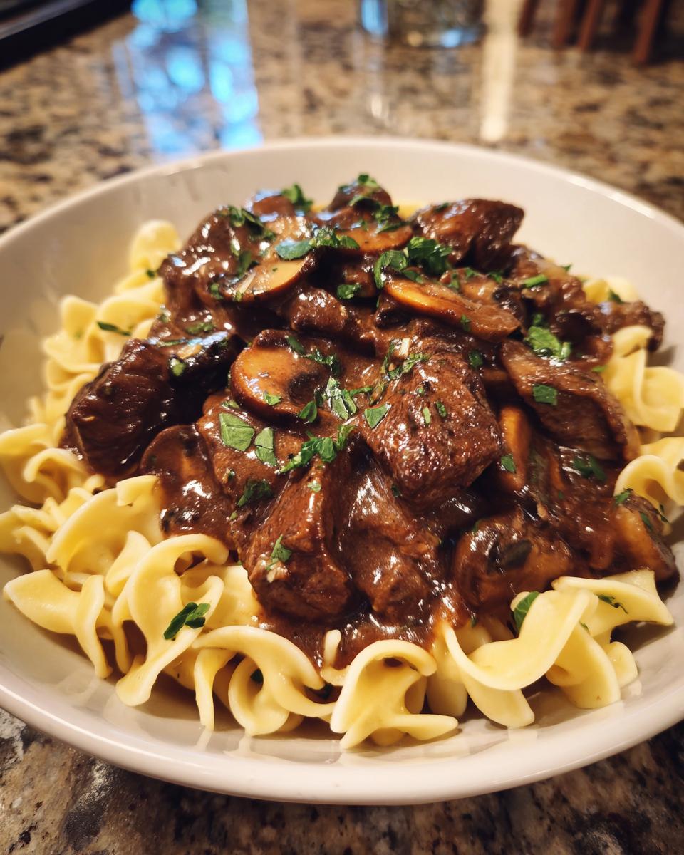 A close-up of a bowl of old fashioned beef stroganoff served over egg noodles, topped with mushrooms and parsley.