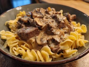 A close-up of a bowl of old fashioned beef stroganoff served over wide egg noodles, featuring tender beef chunks and mushrooms in a creamy sauce.