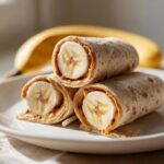 Three peanut butter banana roll-ups stacked on a white plate, with a banana in the background.