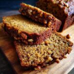 Close-up of moist pumpkin banana bread slices with visible walnuts on a wooden cutting board.
